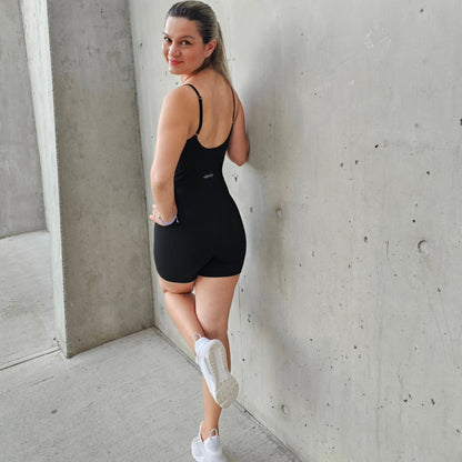 Fitness model leaned against a cement wall posing with short, black, squat proof jumpsuit from Vibras Activewear.